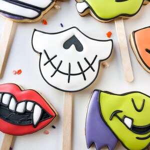 Op de afbeelding: Assortiment van Halloween-koekjes op houten stokjes. De koekjes hebben ontwerpen zoals een mummie, Frankenstein's monster, een schedel, vampierlippen en een groen monster. Versierd met zwarte en witte glazuur en kleurrijke hagelslag.