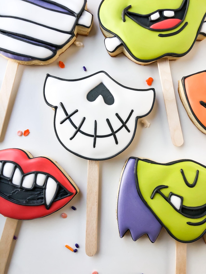 Op de afbeelding: Assortiment van Halloween-koekjes op houten stokjes. De koekjes hebben ontwerpen zoals een mummie, Frankenstein's monster, een schedel, vampierlippen en een groen monster. Versierd met zwarte en witte glazuur en kleurrijke hagelslag.