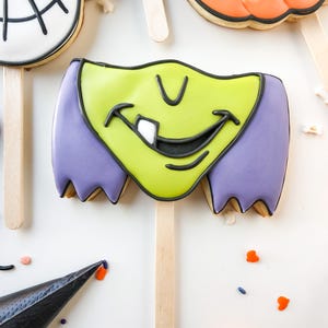 Op de afbeelding: Halloween-thema koekjes op houten stokjes. De koekjes zijn versierd met kleurrijk glazuur, waaronder een heksengezicht met groene huid en paars haar, een pompoen en een schedel. De koekjes zijn gerangschikt op een wit oppervlak met hagelslag en een spuitzak.