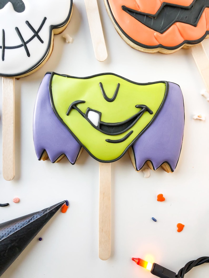 Op de afbeelding: Halloween-thema koekjes op houten stokjes. De koekjes zijn versierd met kleurrijk glazuur, waaronder een heksengezicht met groene huid en paars haar, een pompoen en een schedel. De koekjes zijn gerangschikt op een wit oppervlak met hagelslag en een spuitzak.