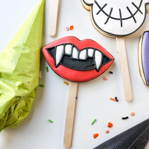 Op de afbeelding: Halloween-koekjes op houten stokjes. De koekjes bevatten een paar rode lippen met hoektanden, een schedel en andere vormen. De koekjes zijn versierd met zwarte, witte en rode glazuur. Strooisels en spuitzakken zijn ook zichtbaar.