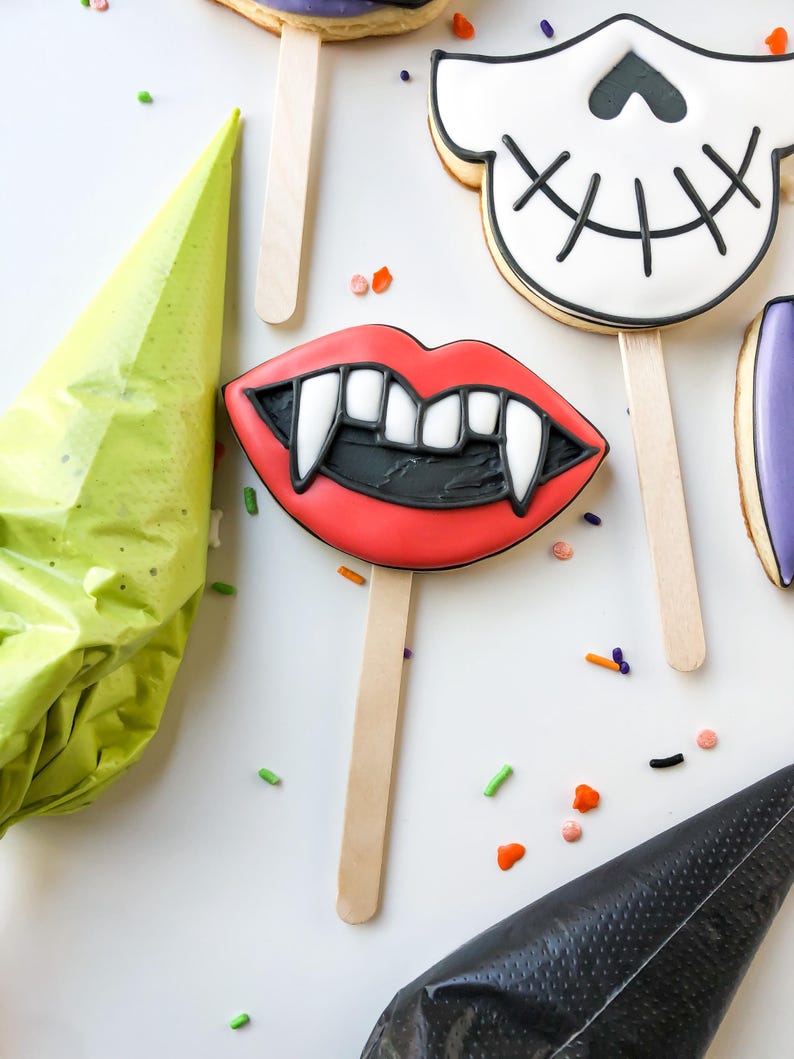 Op de afbeelding: Halloween-koekjes op houten stokjes. De koekjes bevatten een paar rode lippen met hoektanden, een schedel en andere vormen. De koekjes zijn versierd met zwarte, witte en rode glazuur. Strooisels en spuitzakken zijn ook zichtbaar.