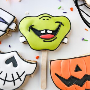 Op de afbeelding: Halloween-thema koekjes op houten stokjes. De koekjes bevatten een groen monstergezicht, een pompoen en een schedel. Elk koekje is versierd met zwarte glazuur en kleurrijke hagelslag. De koekjes zijn gerangschikt op een wit oppervlak.