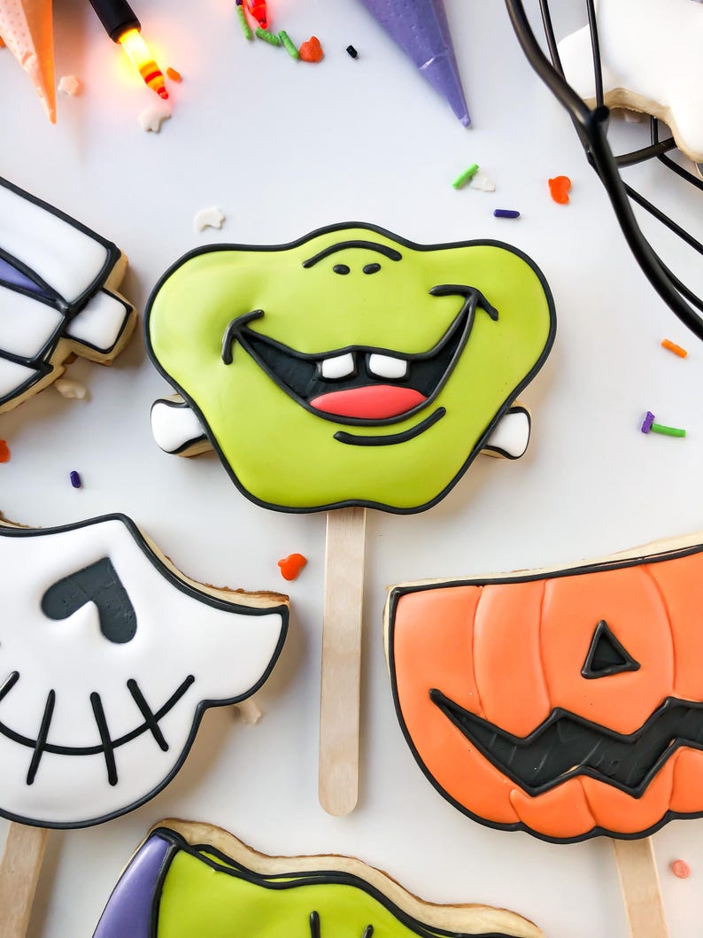 Op de afbeelding: Halloween-thema koekjes op houten stokjes. De koekjes bevatten een groen monstergezicht, een pompoen en een schedel. Elk koekje is versierd met zwarte glazuur en kleurrijke hagelslag. De koekjes zijn gerangschikt op een wit oppervlak.