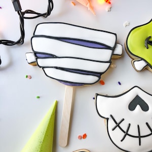Op de afbeelding: Halloween-thema koekjes op stokjes, waaronder een mummie, schedel en groen monster. Het mummiekoekje heeft witte glazuur met zwarte contouren en paarse accenten. Een lichtsnoer en een spuitzak zijn ook aanwezig.