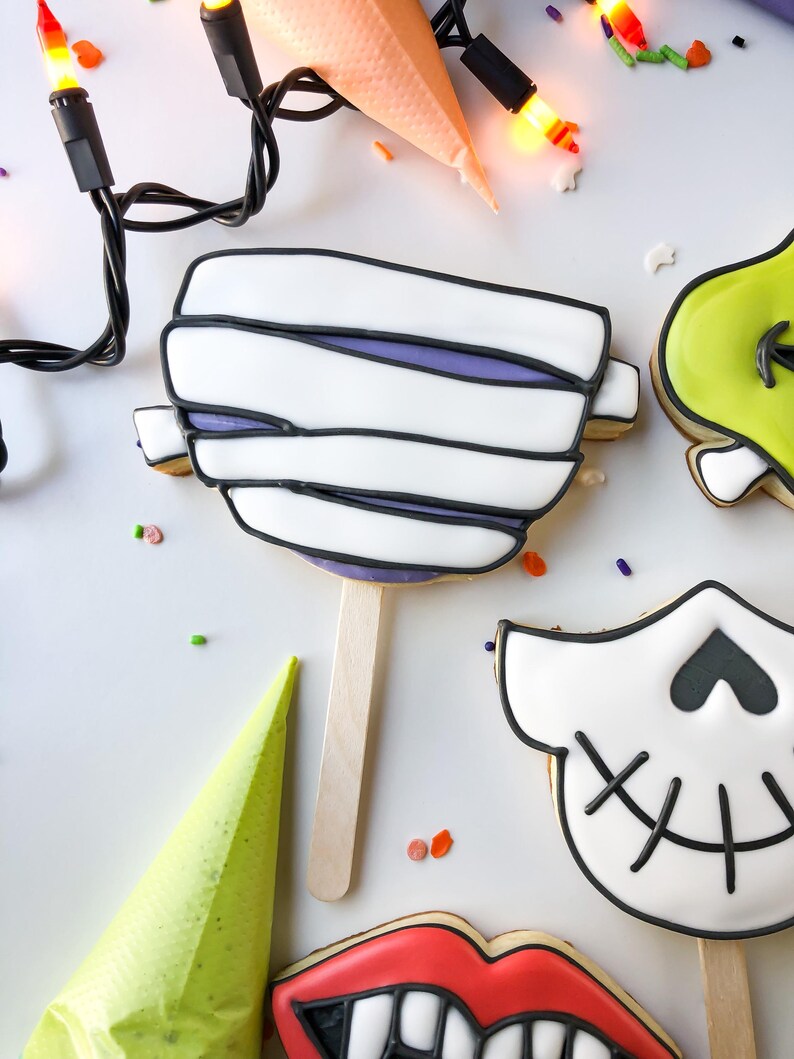 Op de afbeelding: Halloween-thema koekjes op stokjes, waaronder een mummie, schedel en groen monster. Het mummiekoekje heeft witte glazuur met zwarte contouren en paarse accenten. Een lichtsnoer en een spuitzak zijn ook aanwezig.