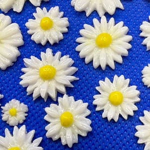 May include: White daisy flower embellishments with yellow centers are scattered on a blue textured surface. The flowers vary in size, with detailed petals. These decorative elements are suitable for crafts and DIY projects.