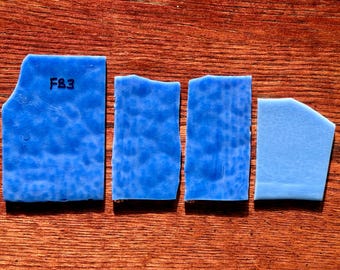 May include: Four irregularly shaped, textured blue glass pieces are arranged on a wooden surface. The largest piece has "F&3" written on it. The pieces vary in shape and shade of blue.