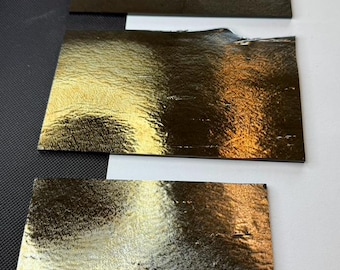 May include: Four rectangular sheets with a highly reflective, metallic gold finish. Each sheet features a textured, crinkled surface that catches the light, creating varied highlights and shadows across the dark, shimmering, and uneven metallic material.