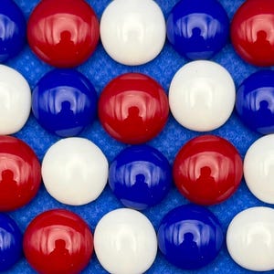 May include: Close-up of a collection of red, white, and blue glass marbles arranged in a grid pattern. The marbles are glossy and reflect light, creating a vibrant display. The background is a textured blue surface.