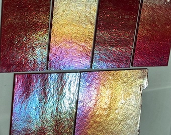 May include: A collection of rectangular glass tiles with a textured, iridescent surface. The tiles display a range of colors, including red, gold, purple, and blue, creating a shimmering effect. The tiles vary in size and shape.