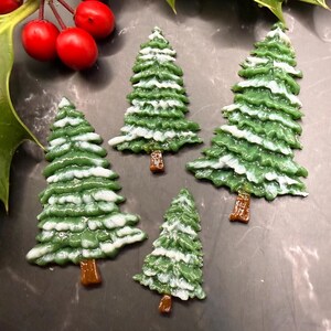May include: Five green Christmas tree ornaments with white snow accents and brown trunks. The ornaments are arranged on a dark surface, with holly leaves and red berries in the background.