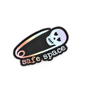 Peut inclure: Un patch thermocollant noir et blanc avec une épingle à nourrice arc-en-ciel et un crâne avec des cœurs pour yeux. Le texte "safe space" est écrit sous l'épingle à nourrice.