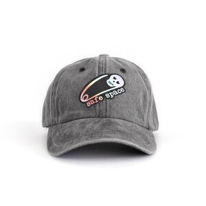 Peut inclure: Une casquette de baseball noire, usée, avec une épingle à nourrice et un crâne en couleur arc-en-ciel. Le texte "safe space" est imprimé sur la casquette.