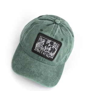 Peut inclure: Une casquette de baseball verte avec un patch noir qui dit "Nature is my religion" et représente une scène de nature avec un papillon, des fleurs et des feuilles.