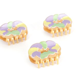 Op de afbeelding: Drie bloemvormige haarclips. Elke clip heeft een goudkleurige basis met tanden en een bloemenpatroon in paars, geel en groen. De clips zijn gerangschikt op een witte achtergrond.