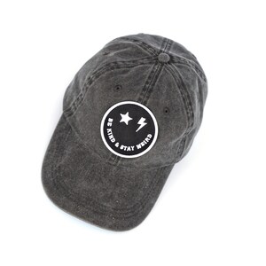 May include: A black denim baseball cap with a black and white embroidered patch that says "Be Kind & Stay Weird" with a smiley face, two stars, and a lightning bolt.