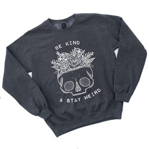 May include: Dark gray sweatshirt with a white skull graphic wearing a floral crown. The text "BE KIND & STAY WEIRD" is printed above and below the skull. The sweatshirt has long sleeves and a crew neck.