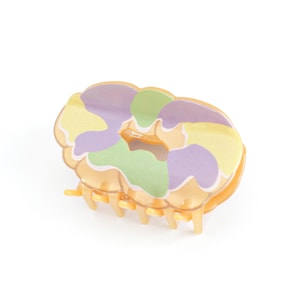 Op de afbeelding: Een decoratieve haarclip in donutvorm. De clip heeft een pastelkleurenpalet van geel, groen en paars. De clip is gemaakt van plastic en heeft een lichtoranje kleur.