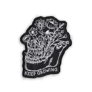 May include: Black embroidered patch featuring a stylized skull with white flowers blooming from within. The patch has the words "KEEP GROWING" in white text at the bottom. The patch has a black border.