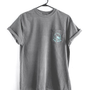 May include: A gray t-shirt with a pocket on the left side. The pocket has a skull graphic and the text "stay weird" in turquoise.