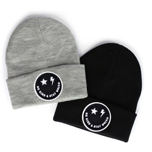 May include: Two knit beanies with a black and white smiley face patch that says "Be Kind & Stay Weird". One beanie is gray and the other is black.