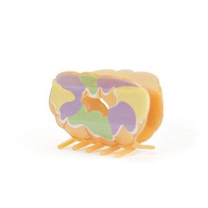 Op de afbeelding: Een kleurrijke haarklauwclip in tinten oranje, geel, groen en paars. De clip heeft een uniek, abstract ontwerp met een afgeronde vorm en meerdere tanden om het haar vast te zetten. De clip is gemaakt van plastic.