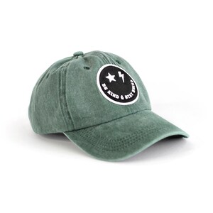 May include: A green baseball cap with a black and white smiley face patch that says "Be Kind & Stay Weird".