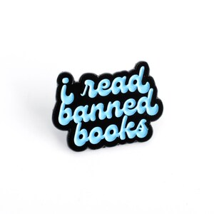 May include: A black enamel pin with the words "i read banned books" in light blue script. The pin has a black border and a white background.