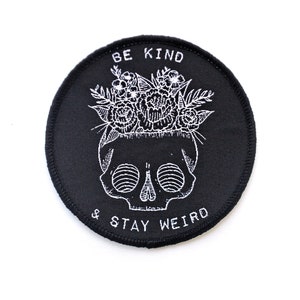 May include: Black and white embroidered patch with a skull wearing a flower crown. The text reads "Be Kind & Stay Weird".