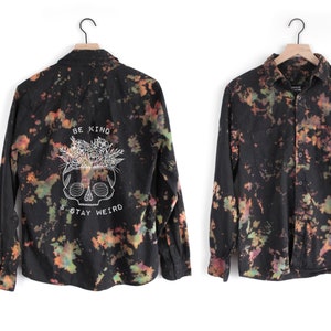May include: Black long-sleeved button-up shirt with a tie-dye pattern in shades of orange, green, and black. The back of the shirt features a white graphic of a skull with flowers and the text "Be Kind & Stay Weird".
