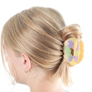 Op de afbeelding: Een blonde haarstijl is in een lage knot gestyled, vastgezet met een kleurrijke haarklem. De klem heeft een mix van pastelkleuren, waaronder geel, paars en groen, wat een speelse touch geeft. Een kleine gouden oorring is zichtbaar.