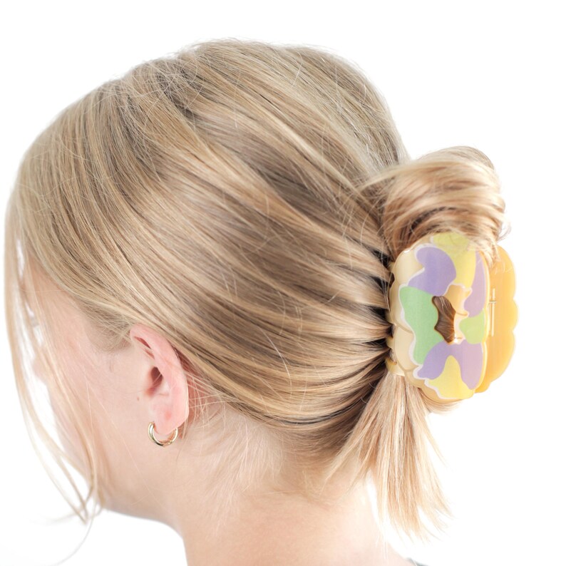 Op de afbeelding: Een blonde haarstijl is in een lage knot gestyled, vastgezet met een kleurrijke haarklem. De klem heeft een mix van pastelkleuren, waaronder geel, paars en groen, wat een speelse touch geeft. Een kleine gouden oorring is zichtbaar.