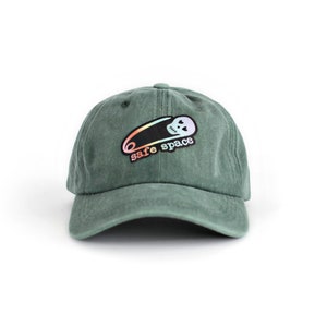 Peut inclure: Une casquette de baseball verte avec une épingle à nourrice arc-en-ciel et un motif de crâne avec le texte "safe space".
