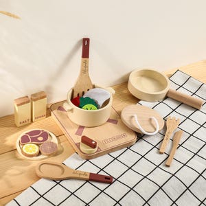 Può includere: Un set da cucina giocattolo in legno con pentola, tagliere e utensili. Il set include un piatto con bistecca, fette di limone e cipolla, più saliera e zuccheriera. Il tagliere ha la scritta "Indoo".