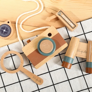 Könnte beinhalten: Holzspielzeug-Set mit Kamera, Fernglas, Kompass, Lupe und einem kleinen Schild mit dem Wort "ADVENTURE". Die Kamera hat eine braune und blaue Linse. Auf der Lupe und dem Fernglas steht der Name "Winifred".