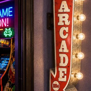 May include: A vertical metal wall sign shaped like an arrow with a distressed red finish and white lettering that reads ARCADE. The sign features five illuminated round light bulbs along the right edge and a circular 25 cents games emblem.