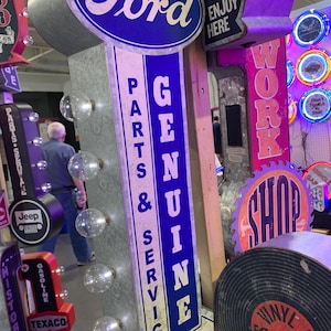 May include: A vintage Ford sign with a blue oval logo and the words "PARTS & SERVICE GENUINE" in white. The sign is an arrow shape with clear light bulbs. Other neon signs are visible in the background.