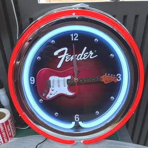 May include: Circular wall clock featuring a red and white Fender Stratocaster guitar design on a black backdrop. The clock has a red neon outer ring and a blue neon inner ring. The Fender logo is displayed at the top.
