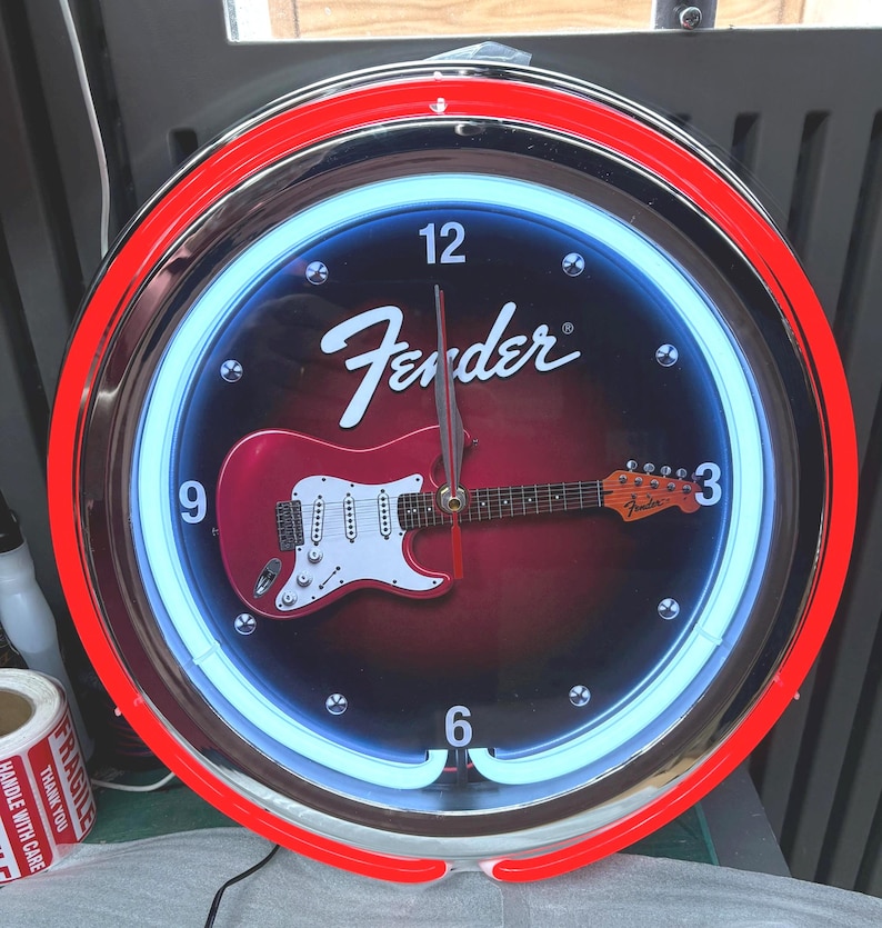 May include: Circular wall clock featuring a red and white Fender Stratocaster guitar design on a black backdrop. The clock has a red neon outer ring and a blue neon inner ring. The Fender logo is displayed at the top.