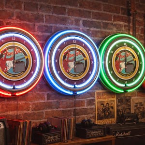 May include: Three Fender neon clocks with red, blue, and green neon rings hang on a brick wall. Each clock face features the Fender logo and a guitar graphic. Guitars and vintage posters are also visible.