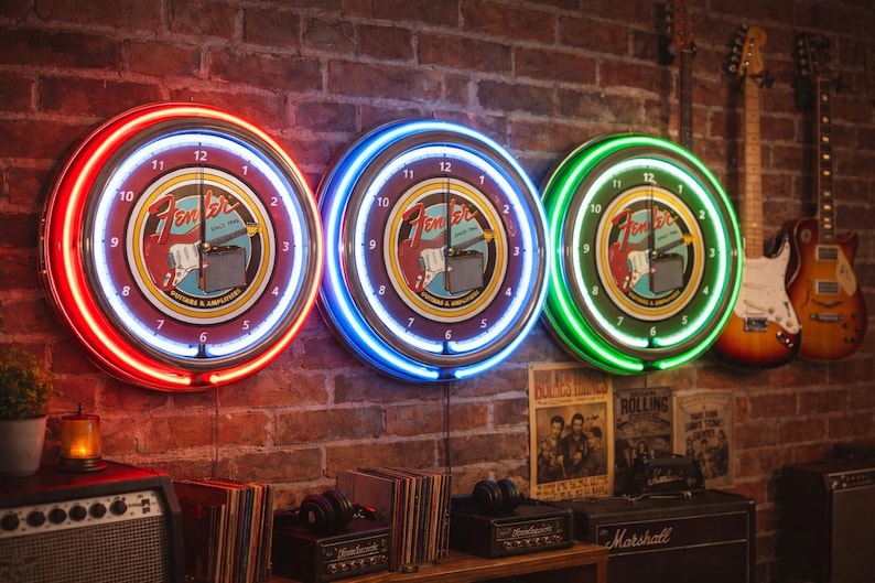 May include: Three Fender neon clocks with red, blue, and green neon rings hang on a brick wall. Each clock face features the Fender logo and a guitar graphic. Guitars and vintage posters are also visible.
