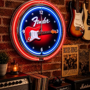 May include: A round Fender neon clock with a red and blue neon border. The clock face features a red guitar graphic and the Fender logo. The clock is mounted on a brick wall, with guitars visible in the background.