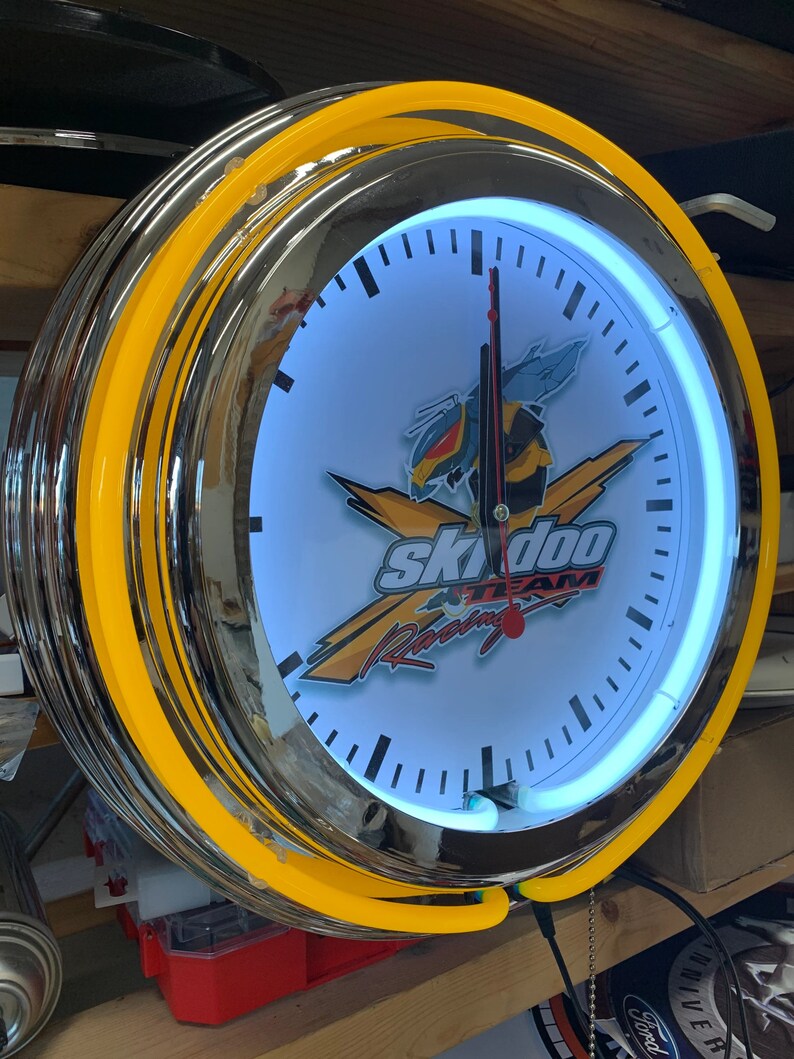 May include: A chrome and yellow neon clock with a white face featuring the Ski-Doo Team Racing logo. The clock has a black and white clock face with black hour and minute hands.