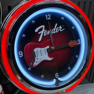 May include: A circular wall clock with a red and chrome surround. The black clock face features a red electric guitar graphic and the Fender logo. A blue neon light encircles the face.