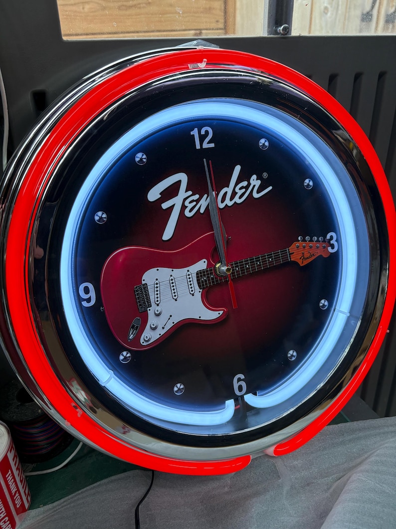 May include: A circular wall clock with a red and chrome surround. The black clock face features a red electric guitar graphic and the Fender logo. A blue neon light encircles the face.
