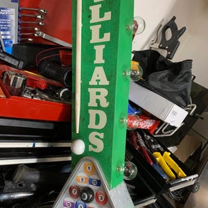 May include: A green sign with the word "BILLIARDS" in white letters. The sign has a triangle of billiard balls at the bottom with a white cue ball and a cue stick on the side.