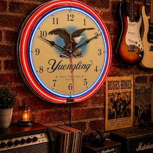 May include: A round wall clock featuring a cream-coloured face with black numerals, a central illustration of an eagle perched on a barrel, and the text Yuengling America's Oldest Brewery Since 1829. The clock is framed by glowing red and blue neon tubes.