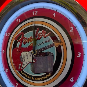 May include: A circular clock with a red and white face, displaying the Fender logo, a red guitar, and an amplifier. The clock has a neon light border and the text "SINCE 1946" and "GUITARS & AMPLIFIERS".