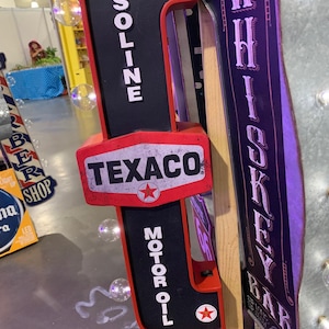 May include: A red and black vintage-style Texaco gas station sign with the words "GASOLINE" and "MOTOR OIL" in white lettering. The sign also features a red star and the word "TEXACO" in white lettering. The sign is mounted on a wooden post.